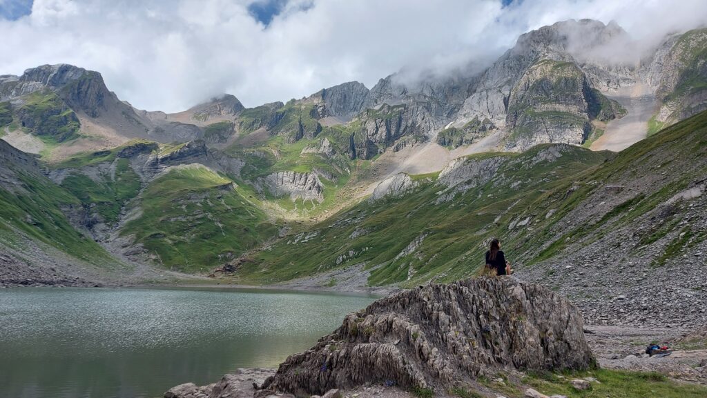 Cindy, votre coach, assise de dos sur un gros rocher au bord d’un lac, faisant face à un massif montagneux, dans un paysage naturel calme